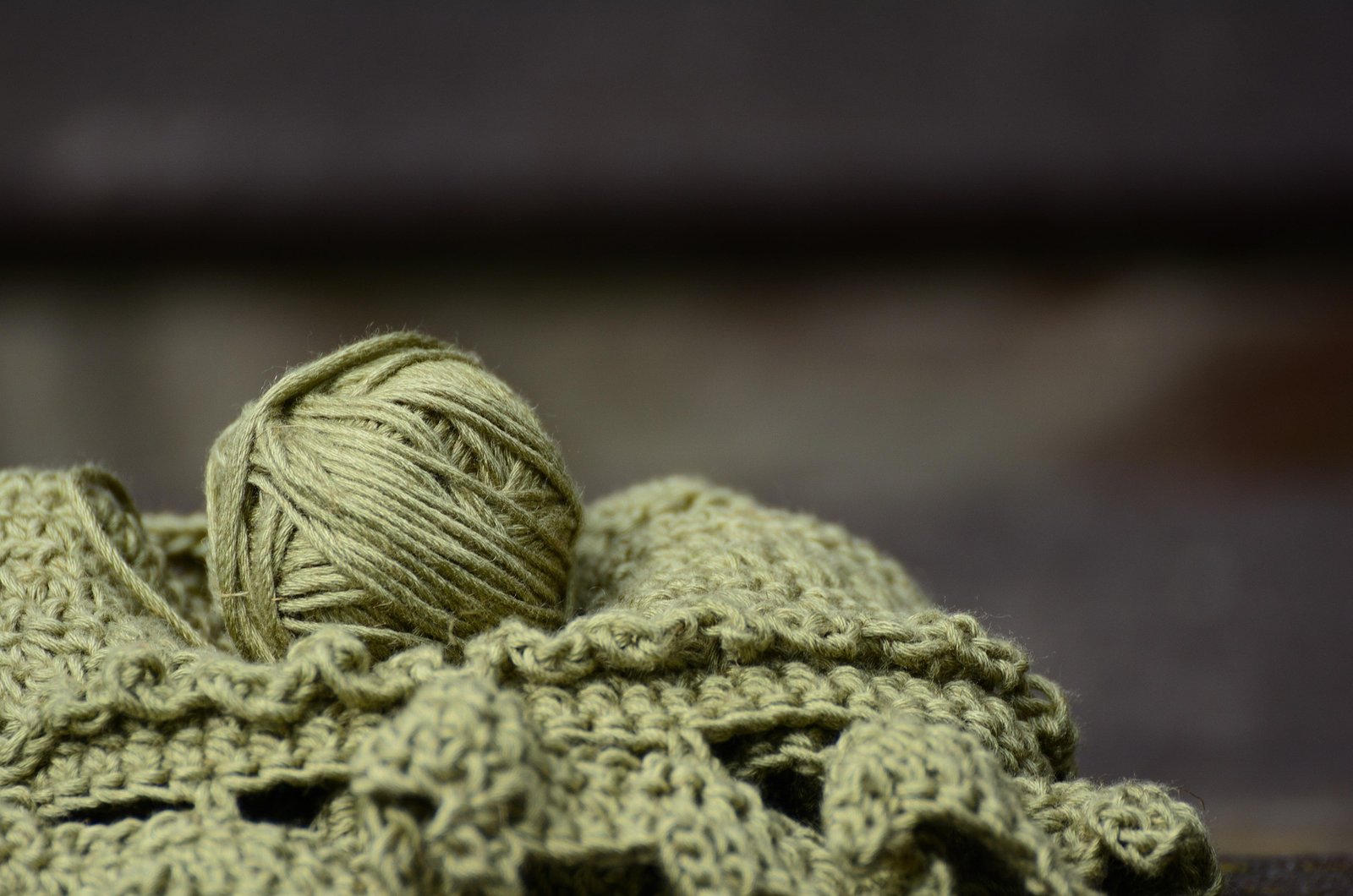 A ball of olive-green yarn rests on a worn wooden surface, with a loose strand trailing toward a tangled pile of yarn behind it.