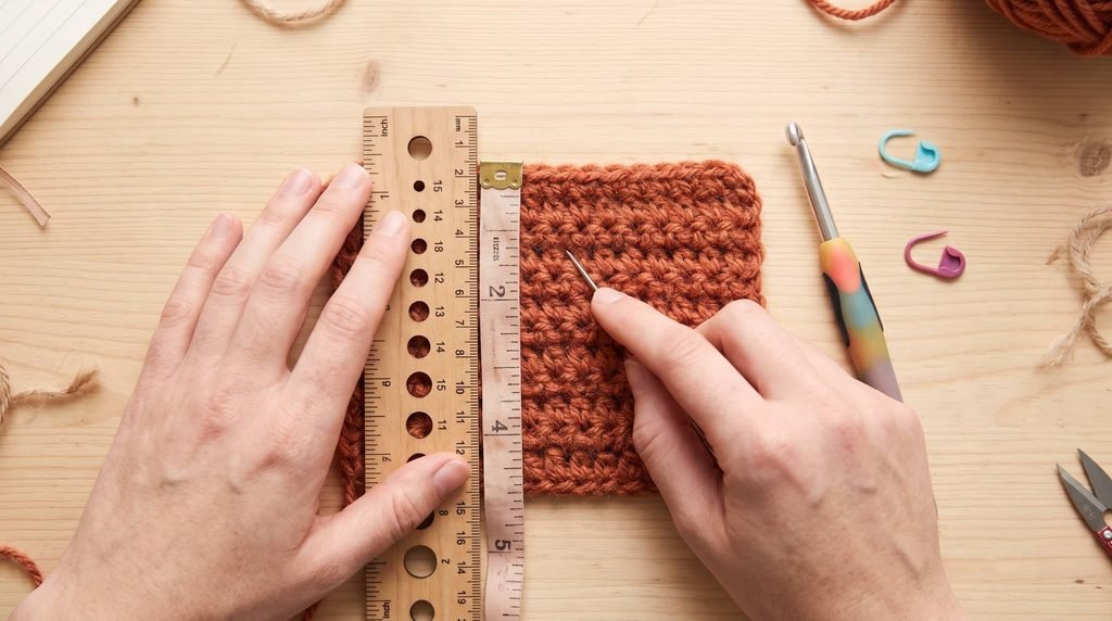Hands measure a rust-colored crochet swatch with a ruler while holding a yarn needle; a crochet hook and stitch markers lie nearby.(cardigans crochet patterns)
